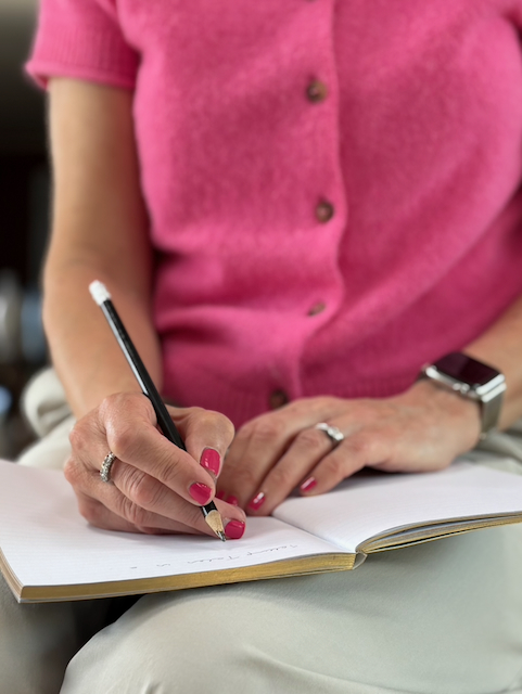 hands of woman wearing pink top writing in a notebook with a pencil